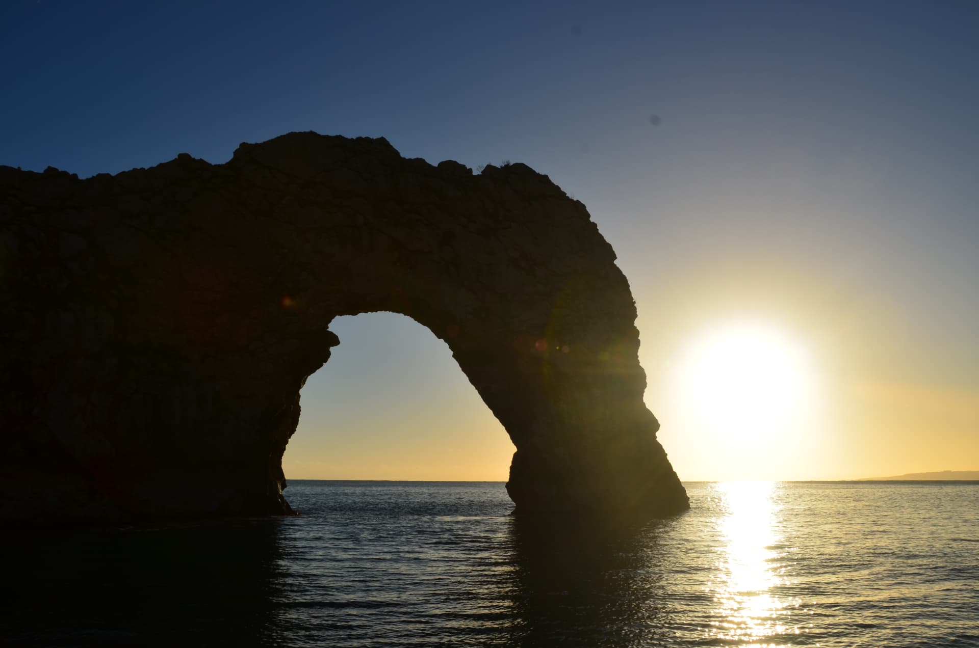A coastal rock arch silhouetted against the setting sun over a calm sea