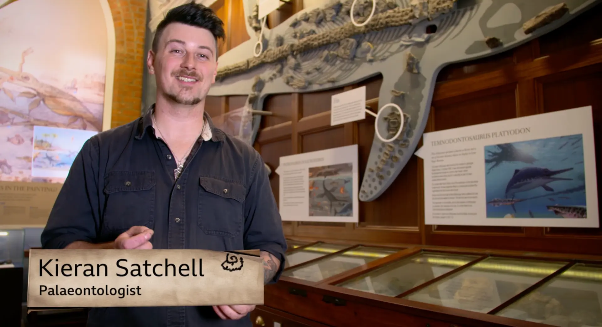 Palaeontologist Kieran Satchell in a museum gallery in front of a large fossilised marine reptile skeleton